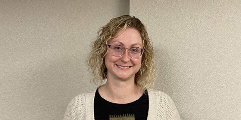 A female with medium length curly blonde hair, wearing glasses, black shirt, beige cardigan, and holding an award.