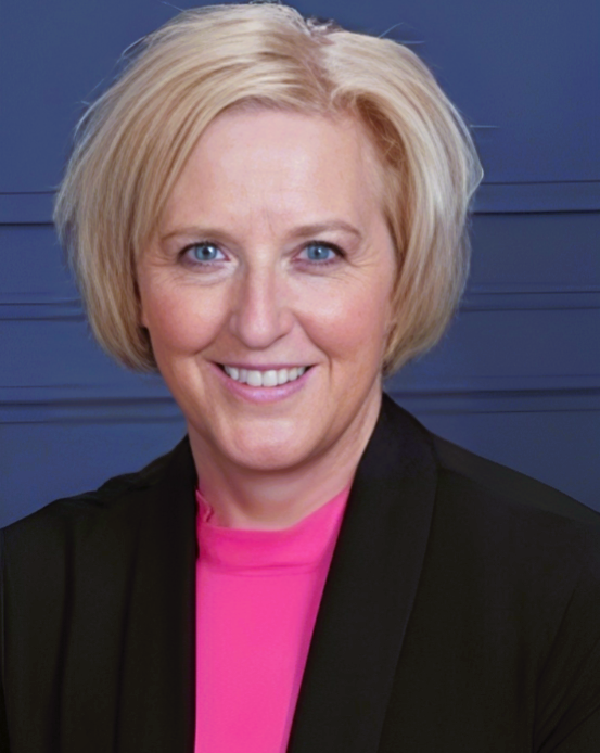 Headshot of a woman with short blonde hair and blue eyes smiling. She is wearing a pink shirt with a black blazer