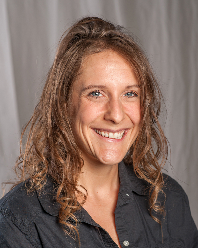 Headshot of a woman with curly medium length brown hair, wearing a dark grey shirt smiling