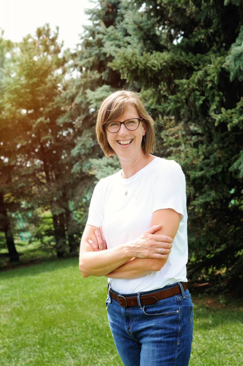 woman with short brown hair and glasses smiling surrounded by green plants. She is wearing a white shirt and blue jeans.