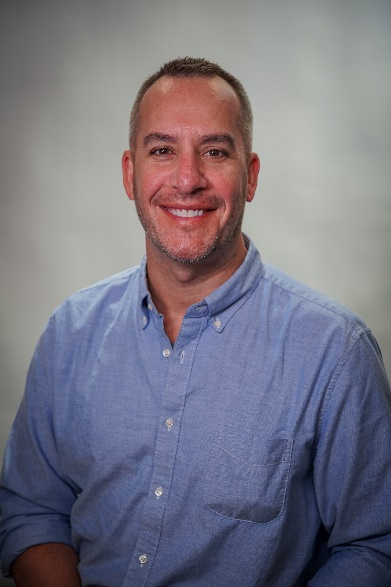 Man with short buzzed hair and facial hair smiling. He is wearing a light blue button up shirt.