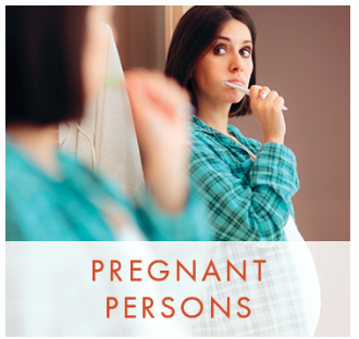 A pregnant woman brushing her teeth and looking at herself in the mirror. 