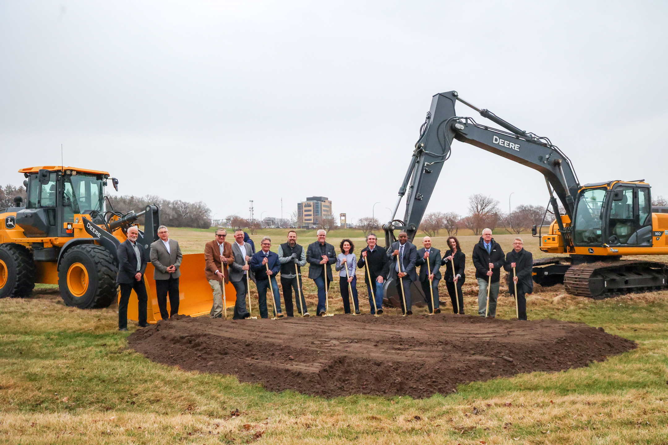 A group of government officials stand ready with their shovels in the ground at the state lab construction site. Two large digging vehicles are behind them.
