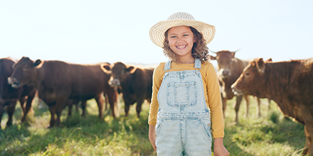 A young girl wearing a hat and overalls stands in front of cows, smiling