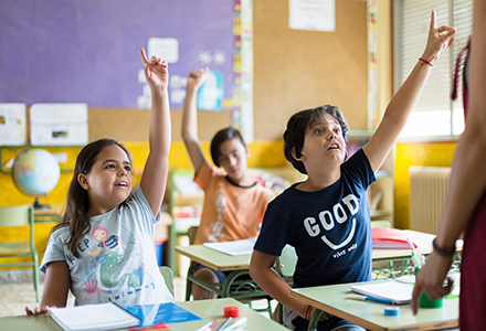 Two students raise their hands eagerly in class. Another student raises his hand behind them.