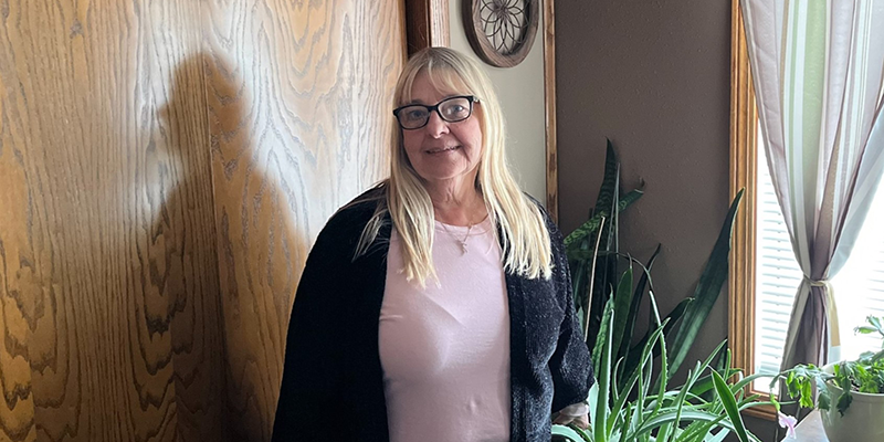 A light skinned female with blond hair and glasses. She is wearing a pink shirt and a black cardigan.