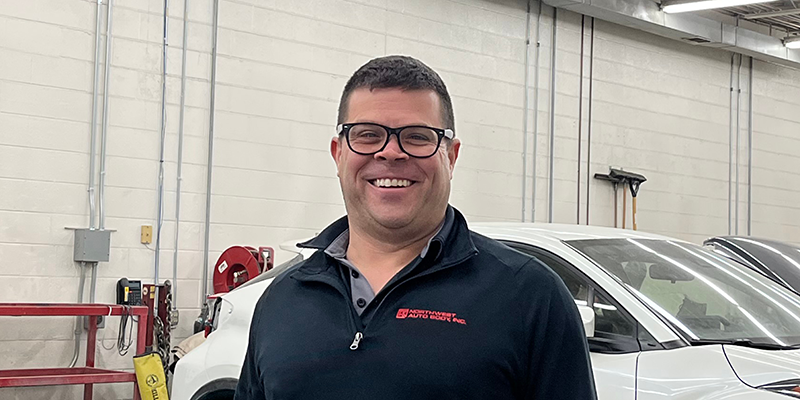 Light skinned middle aged male with short brown hair, glass, a black quarter zip shirt, and khakis. He is standing in a shop with a white car on a hoist.