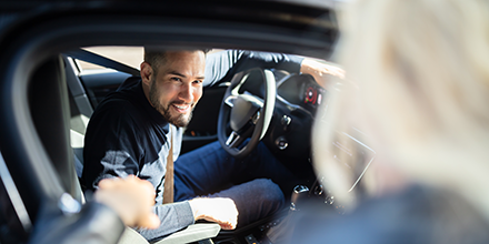 A man in a car, smiling at a lady getting in for a ride.