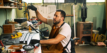 A man in a wheelchair and work apron, working in a shop with many tools laid out on the table in front of him.