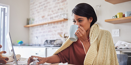 Woman sipping coffee in her kitchen with a blanket around her, on her computer.