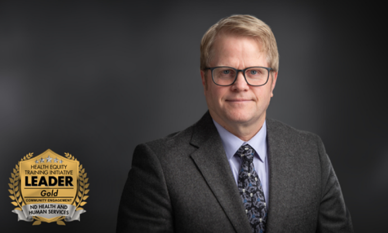 Middle aged white man with glasses and short blonde hair wearing a dark grey suit with a charcoal grey blurred background with a gold health equity recognition badge in the bottom left corner