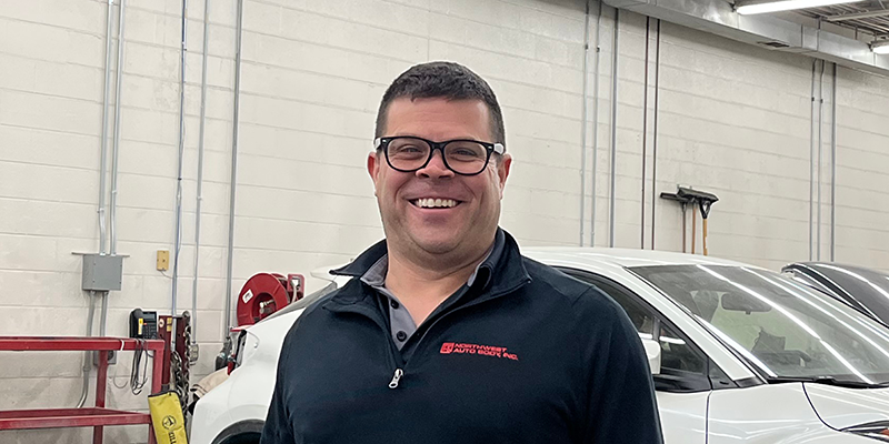  A light skinned middle-aged male with short brown hair, glasses, a black quarter zip shirt, and khakis. He is standing in the garage of the auto body shop next to a black truck with the passenger door open.