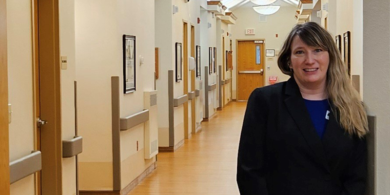 Woman in a black jacket standing in front of a long hallway at a nursing facility  